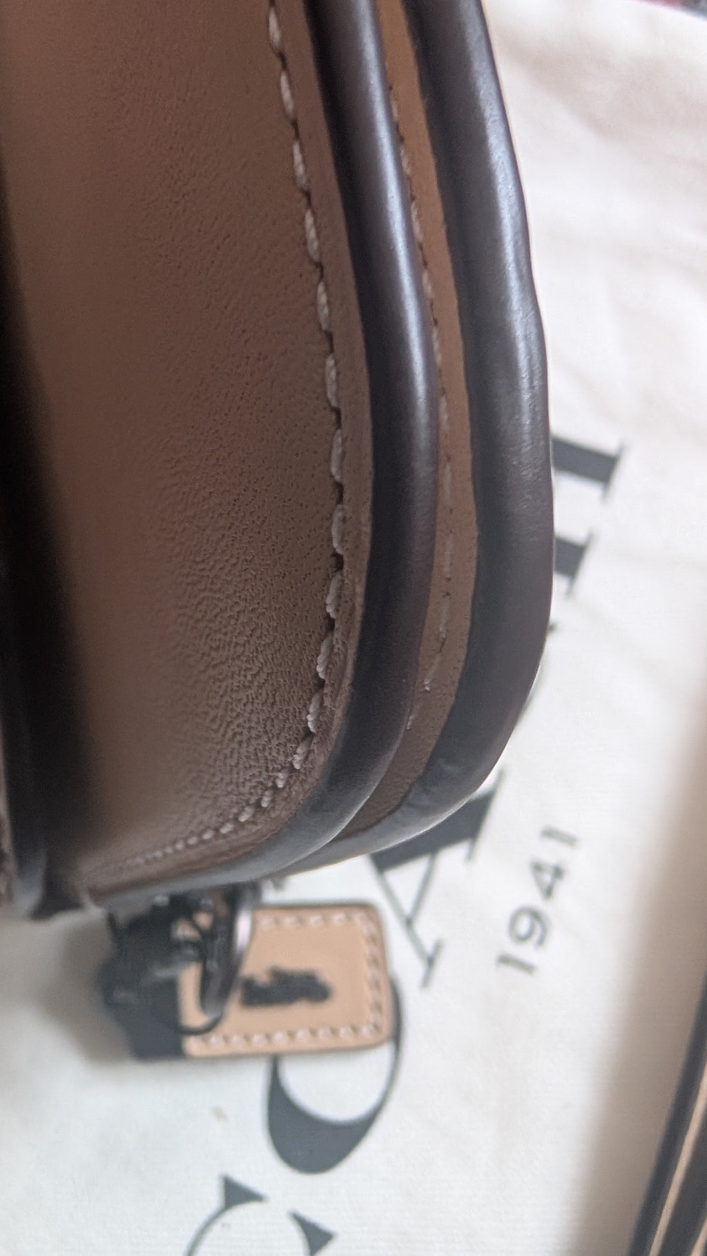 Close-up of a brown leather handbag corner showing the stitching, edge profile, and a small branded tag on a light patterned surface. Pre-Owned Coach Courier Wristlet with Floral Appliqué – Glovetanned Leather