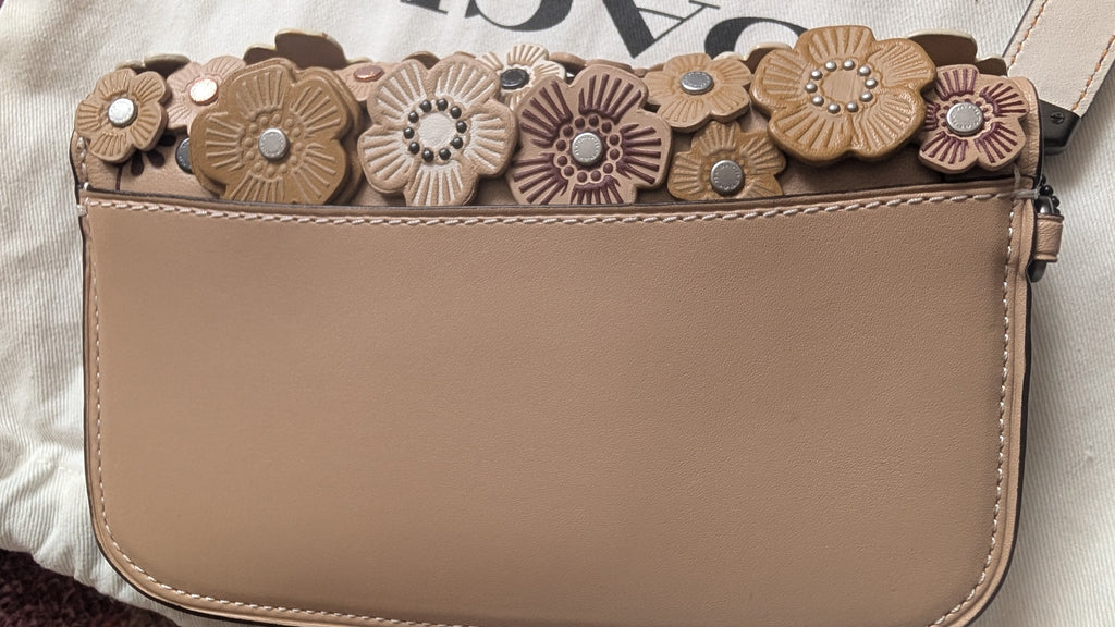 A close-up of a beige leather crossbody bag featuring a decorative side panel adorned with layered faux leather flower appliqués in shades of tan, brown, and dusty rose, accented with small metallic studs. Pre-Owned Coach Courier Wristlet with Floral Appliqué – Glovetanned Leather