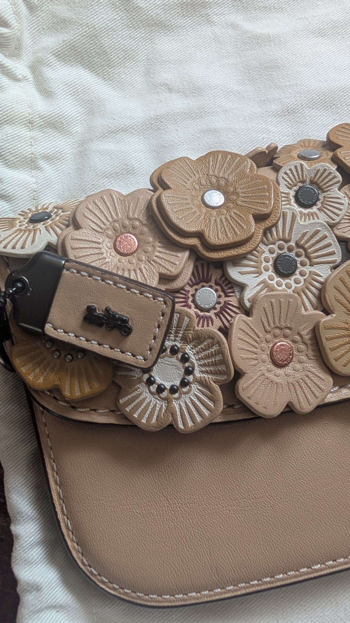 A close-up of a beige leather handbag with a decorative flap covered in 3D flower appliqués in beige, tan, and brown tones, with a matching leather key fob resting on the side. Pre-Owned Coach Courier Wristlet with Floral Appliqué – Glovetanned Leather
