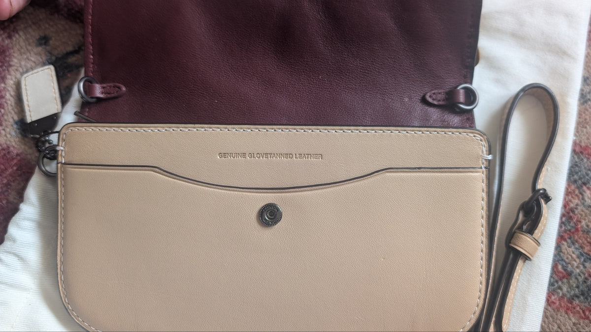 A Vuitton shoulder bag in two-tone leather, featuring a dark burgundy side panel and a beige front flap with a curved line detail and snap button closure. The bag is shown open, revealing its interior, with a white dust bag underneath and a hand holding the bottom left corner. Pre-Owned Coach Courier Wristlet with Floral Appliqué – Glovetanned Leather