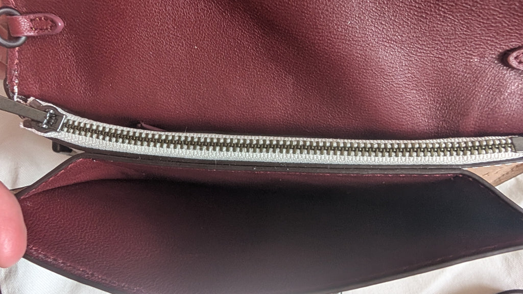 Close-up of a burgundy leather handbag showing the interior lining, a silver zipper, and a finger holding the flap open. Pre-Owned Coach Courier Wristlet with Floral Appliqué – Glovetanned Leather