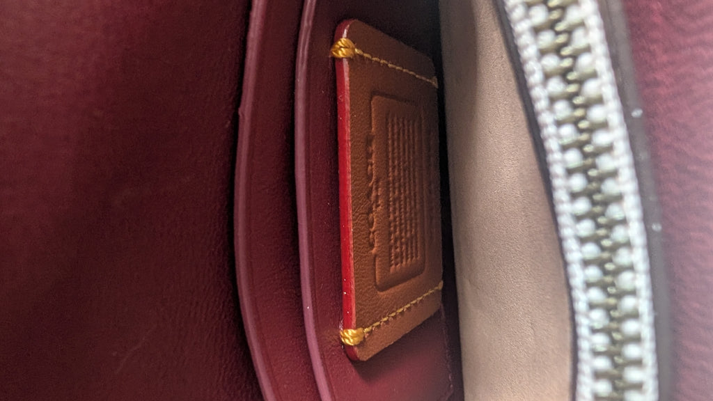 Close-up of the interior of a burgundy leather bag, showing a brown leather insert card with gold stitching and a silver zipper track. Pre-Owned Coach Courier Wristlet with Floral Appliqué – Glovetanned Leather