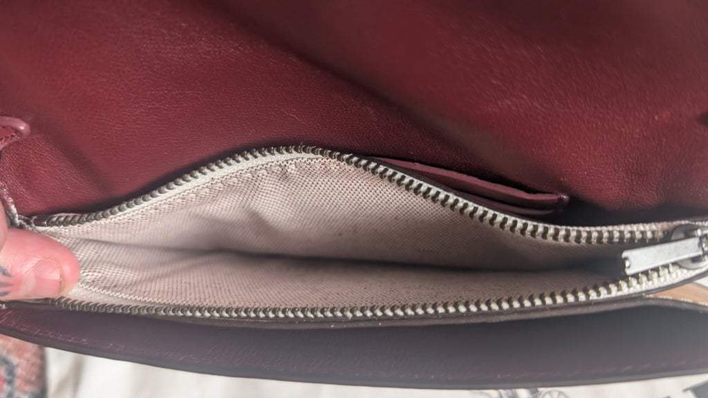 Close-up of a burgundy leather bag interior showing a silver zipper and light beige fabric lining, with a finger holding the edge open. Pre-Owned Coach Courier Wristlet with Floral Appliqué – Glovetanned Leather