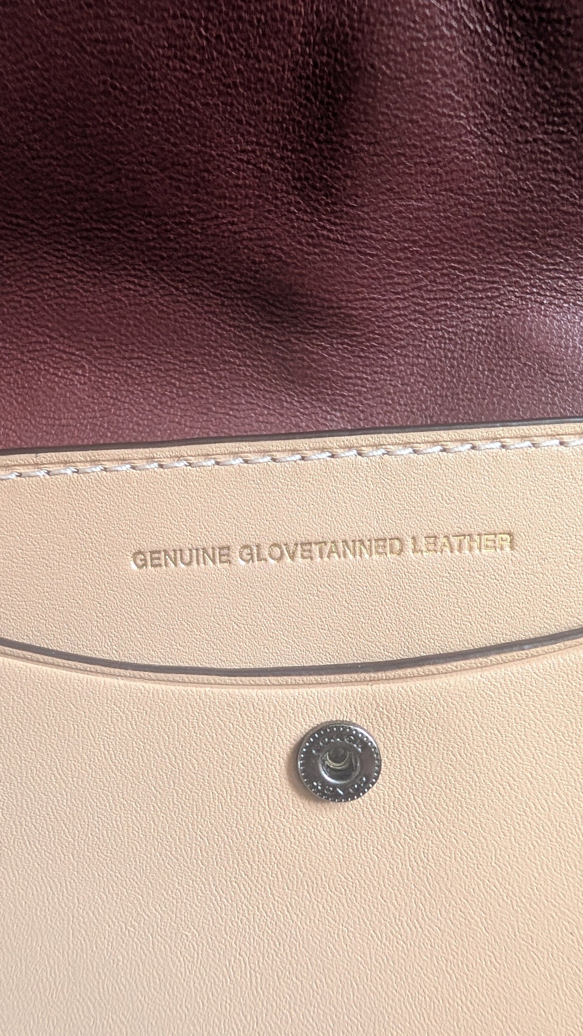 Close-up detail of a leather bag showing a deep burgundy leather panel, a beige leather panel with white stitching, and a silver-tone metal snap button. The text 'GENUINE CLOTH LEATHER' is embossed vertically. Pre-Owned Coach Courier Wristlet with Floral Appliqué – Glovetanned Leather