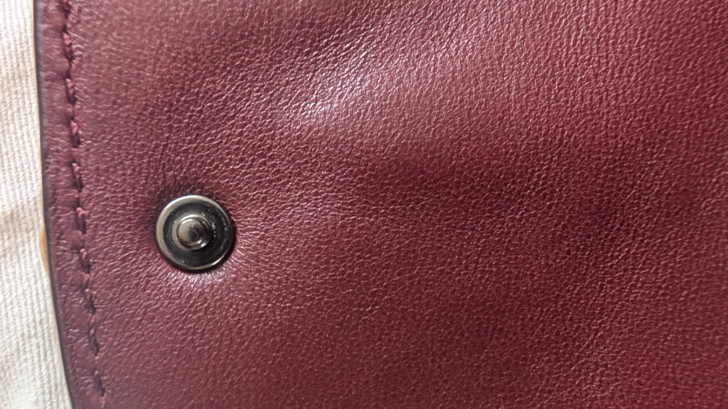 Close-up of a burgundy leather surface with a dark metal snap button and visible stitching along the left edge. Pre-Owned Coach Courier Wristlet with Floral Appliqué – Glovetanned Leather