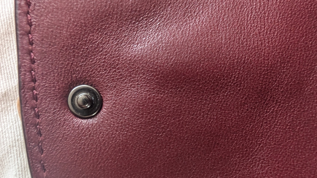 Close-up of a burgundy leather surface with a dark metal snap button and visible stitching along the left edge. Pre-Owned Coach Courier Wristlet with Floral Appliqué – Glovetanned Leather