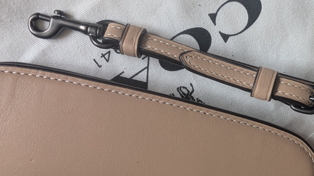 Close-up of a beige leather strap with a silver-toned metal clasp, laid over a matching beige pouch or case on a white dust bag with black logo print and a small 'New Again' tag. Pre-Owned Coach Courier Wristlet with Floral Appliqué – Glovetanned Leather