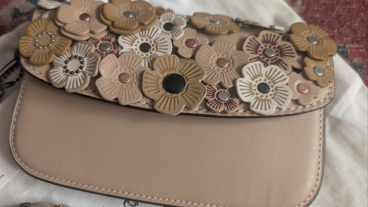 A beige leather shoulder bag with a flap covered in 3D floral appliqués in shades of tan, brown, and cream. The bag has a silver-tone snap closure and is shown with a white 'New Again' brand tag on a white fabric background. Pre-Owned Coach Courier Wristlet with Floral Appliqué – Glovetanned Leather