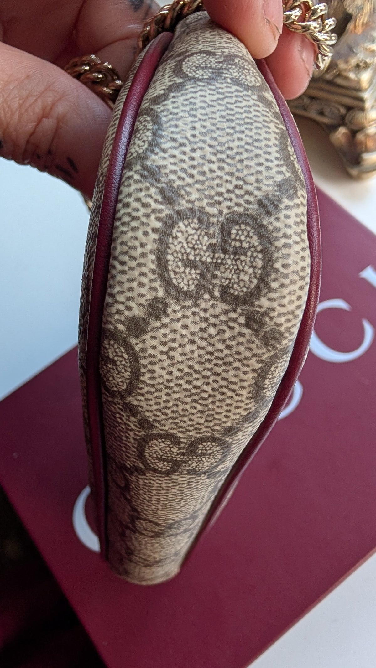 A close-up of a hand holding a Gucci monogram canvas pouch or clutch with dark red leather trim and a gold-tone chain, positioned over a red Gucci box. Pre-Owned Gucci GG Supreme Ophidia Panda Mini Chain Shoulder Bag