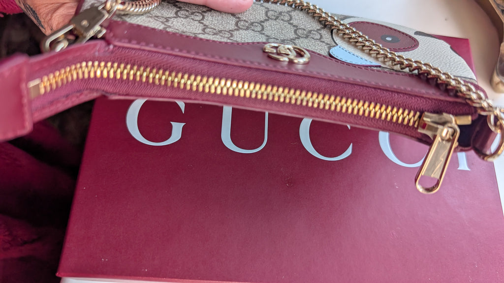 A close-up of a Gucci wristlet pouch in burgundy leather and GG Supreme canvas, featuring gold-tone hardware and a chain strap, resting on a burgundy Gucci box. Pre-Owned Gucci GG Supreme Ophidia Panda Mini Chain Shoulder Bag