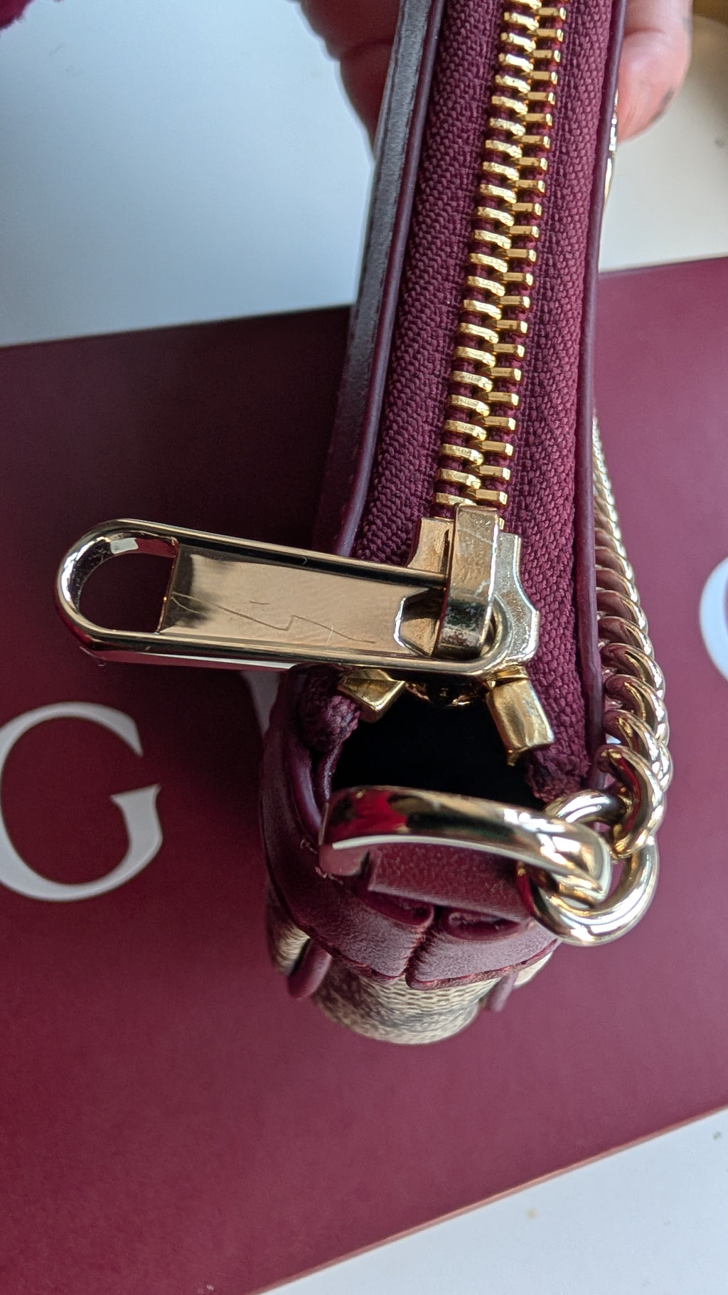 A close-up of a burgundy leather handbag with gold-tone hardware, showing a zipper closure and a gold chain link detail, held by a hand over a burgundy and white surface. Pre-Owned Gucci GG Supreme Ophidia Panda Mini Chain Shoulder Bag