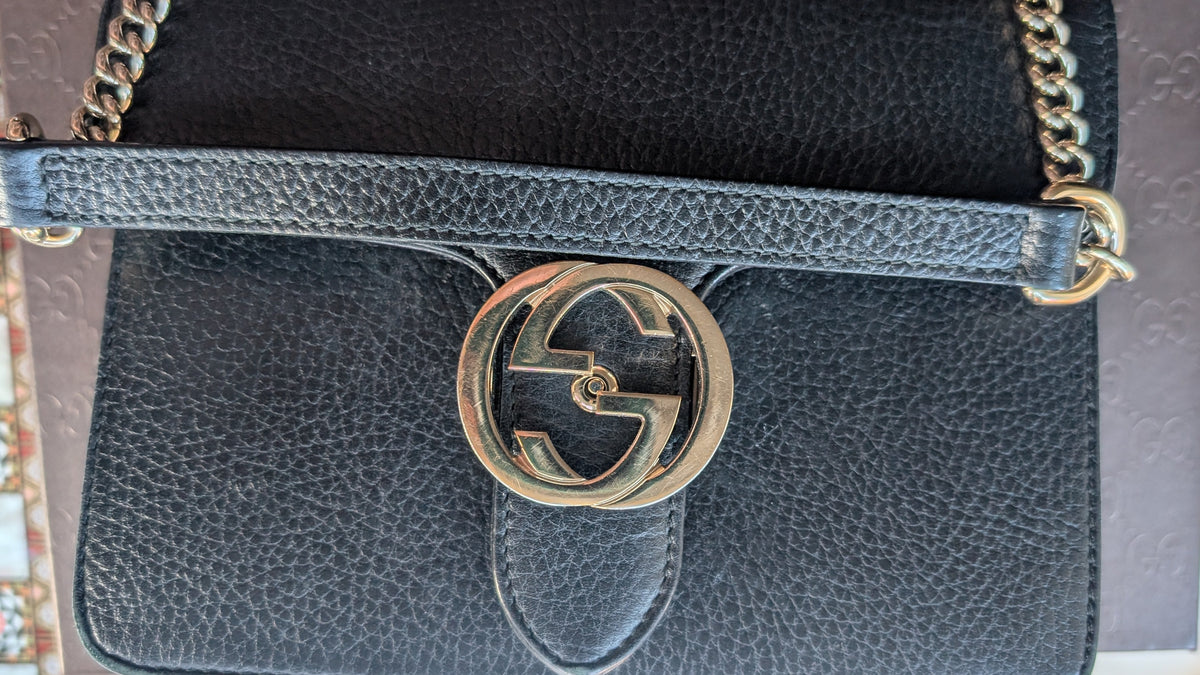 A close-up of a black pebbled leather shoulder bag with a gold-tone interlocking G logo clasp and a matching black leather strap, resting on a brown Gucci box. Pre-Owned Gucci Interlocking Black shoulder bag 