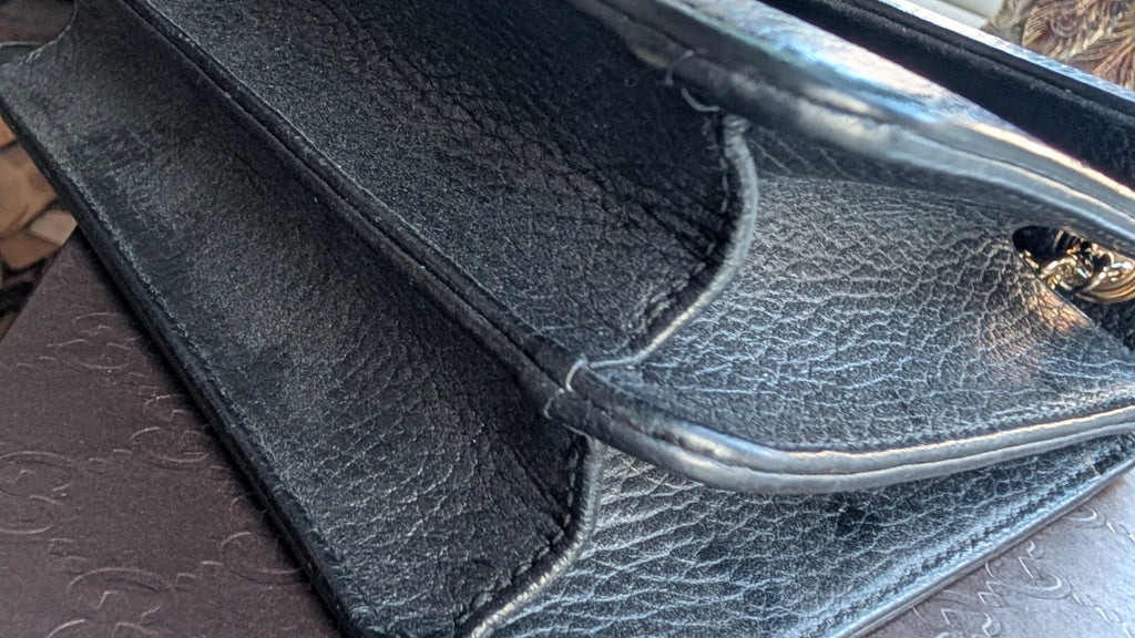 A close-up of a black pebbled leather clutch or handbag corner with a shiny gold tone metal handle, resting on a dark brown textured surface. Pre-Owned Gucci Interlocking Black shoulder bag 