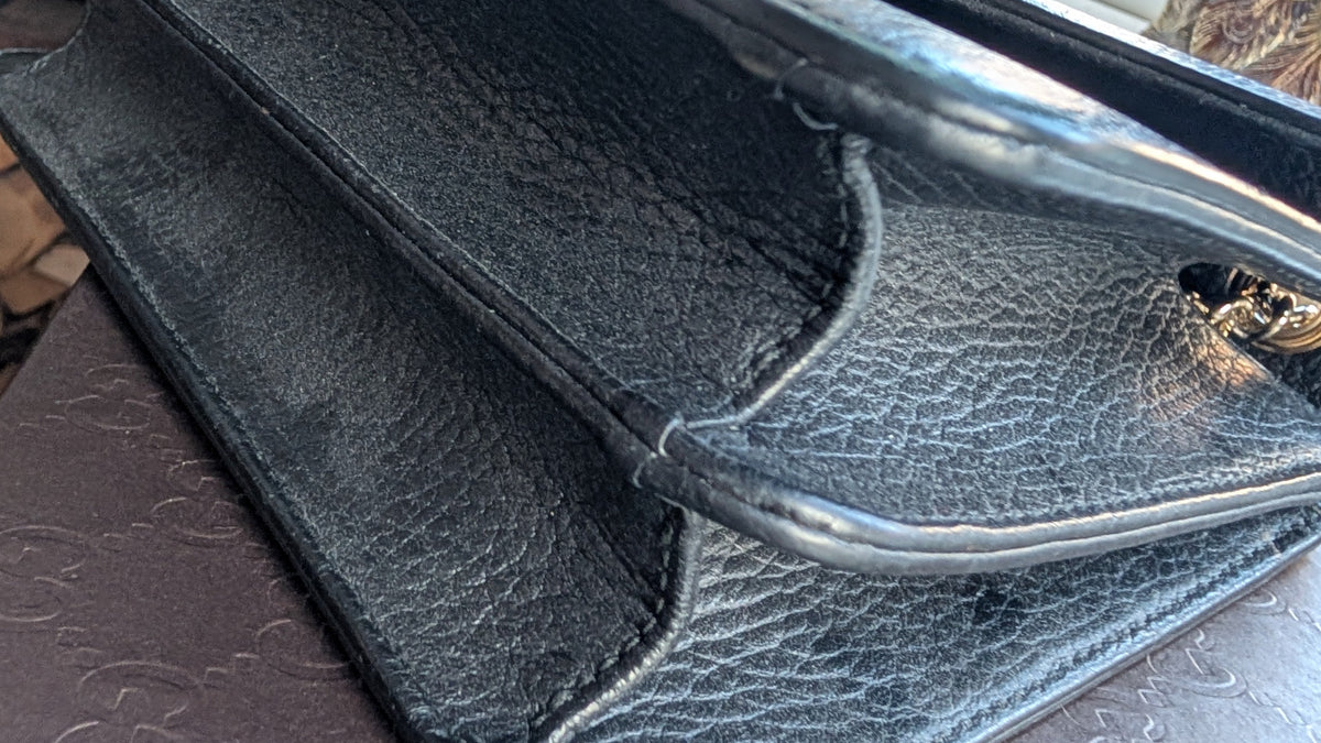 A close-up of a black pebbled leather clutch or handbag corner with a shiny gold tone metal handle, resting on a dark brown textured surface. Pre-Owned Gucci Interlocking Black shoulder bag 