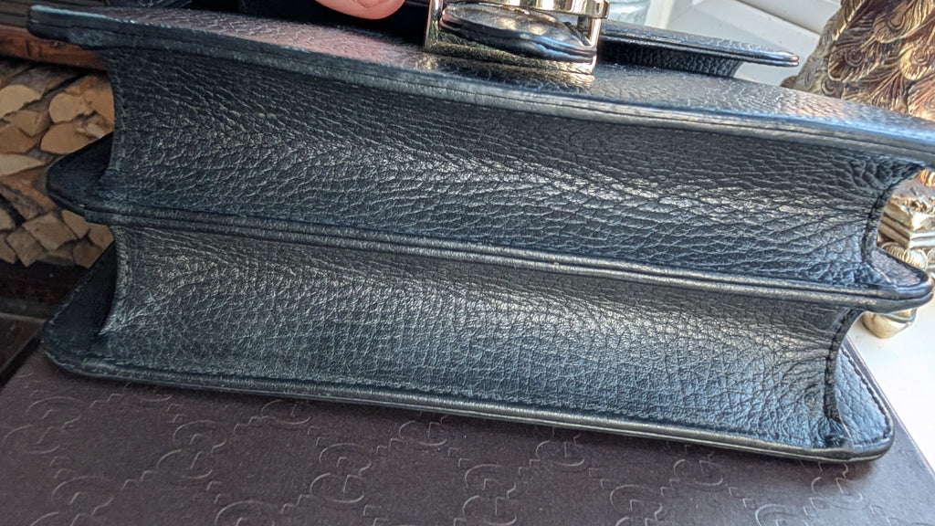 A close-up of a black pebbled leather book-style clutch or small handbag, showing the thick textured spine and a metallic clasp on top, resting on a patterned surface. Pre-Owned Gucci Interlocking Black shoulder bag 