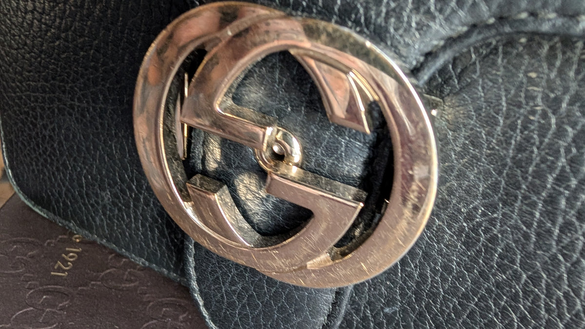 Close-up of a metallic interlocking G logo buckle on a black textured leather , with part of a matching embossed leather card visible underneath. Pre-Owned Gucci Interlocking Black shoulder bag 