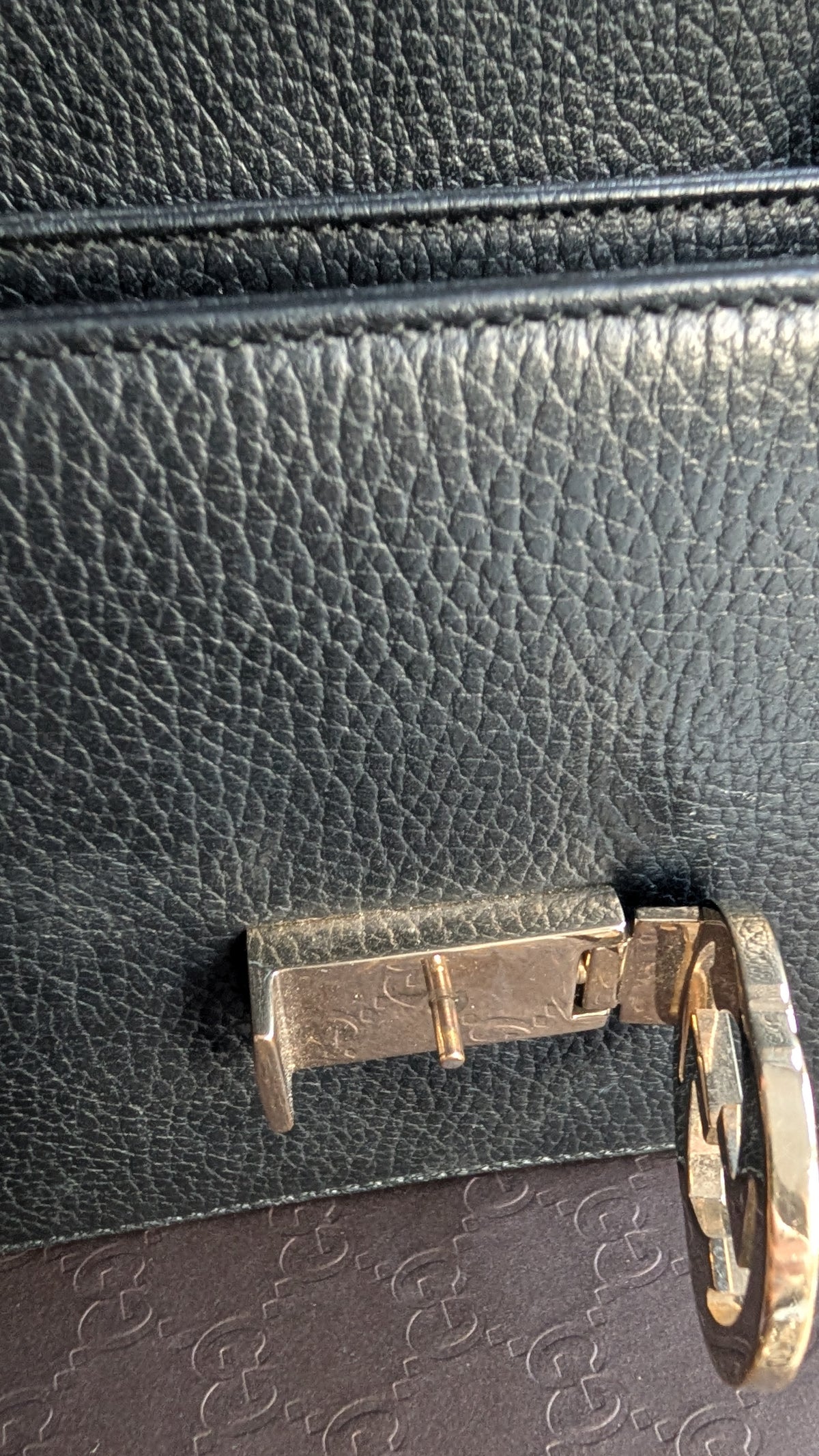 Close-up of a black pebbled leather bag with a gold-tone metal lock and a circular ring detail, resting on a dark brown patterned surface. Pre-Owned Gucci Interlocking Black shoulder bag 