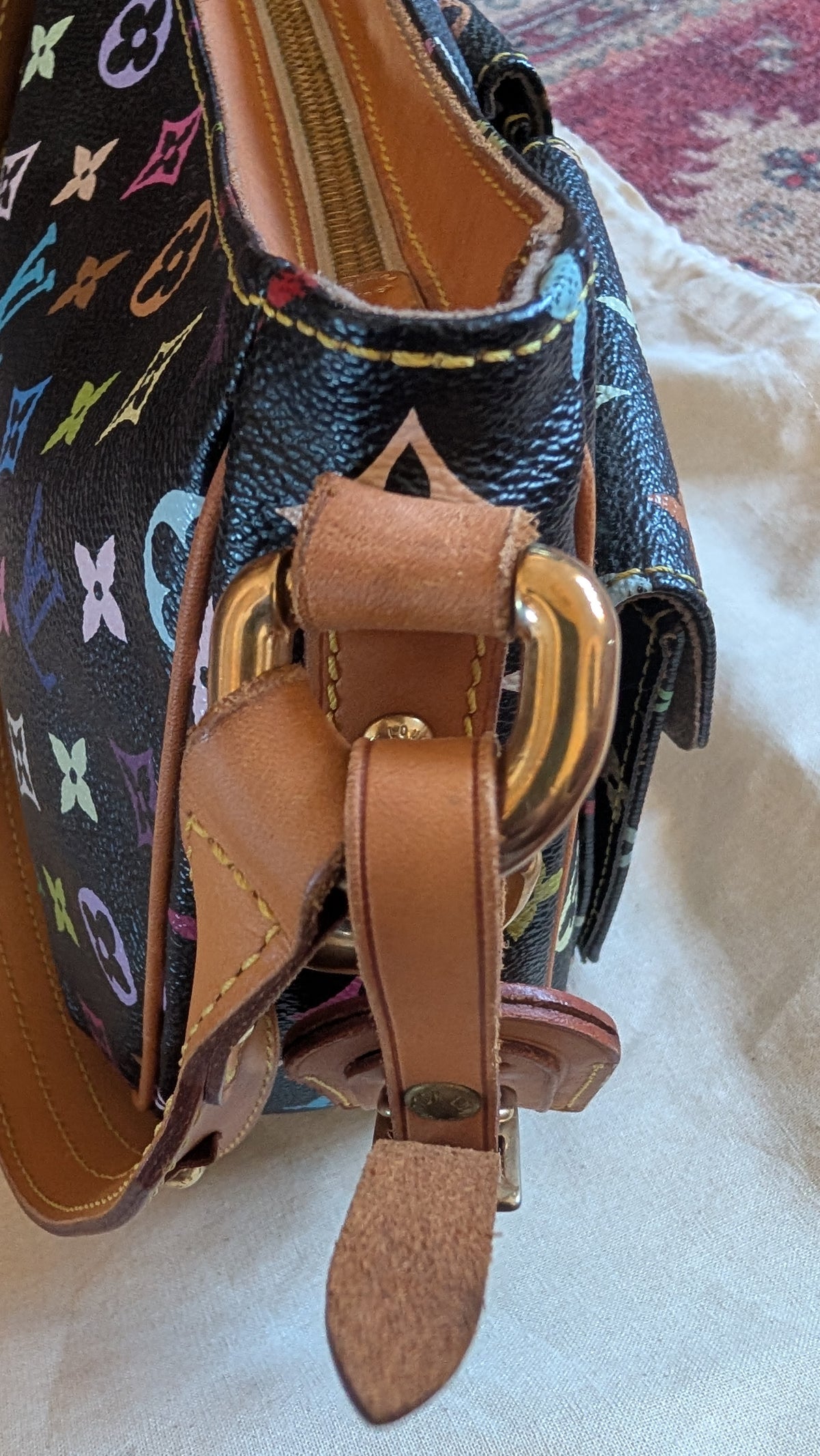 Close-up of a Louis Vuitton shoulder bag in black monogram canvas with colorful LV logos, tan leather straps, and gold-tone hardware, resting on a light fabric surface. Pre-Owned Louis Vuitton Takashi Murakami Lodge PM Shoulder Bag
