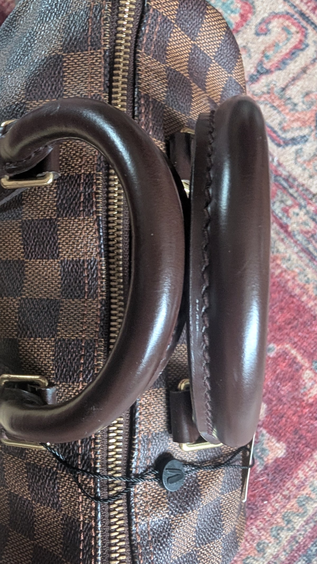 A close-up of a brown leather handle and zipper area of a Louis Vuitton bag in the Damier Ebene checkered pattern, resting on a patterned red and blue rug.Pre-Owned Louis Vuitton Speedy 25 in classic Damier Ebene coated canvas
