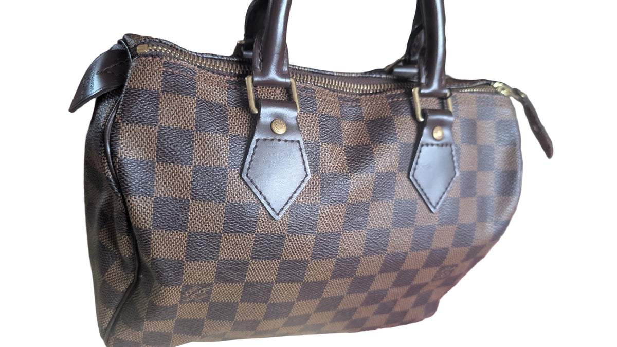 A Louis Vuitton Damier Ebene canvas handbag with brown leather handles and trim, resting on a beige surface over a patterned rug. Pre-Owned Louis Vuitton Speedy 25 in classic Damier Ebene coated canvas
