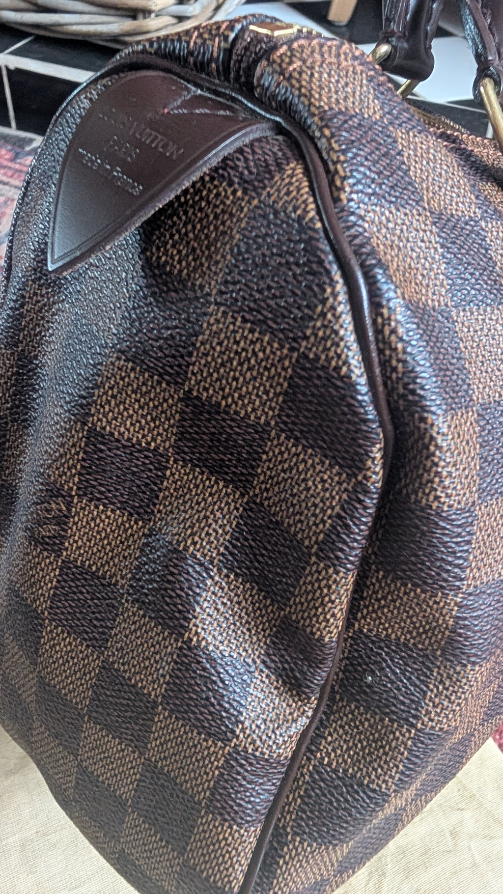 A close-up of a Louis Vuitton handbag in the Damier Ebene checkered canvas, showing the brown and dark brown patterned material and a brown leather logo patch. Pre-Owned Louis Vuitton Speedy 25 in classic Damier Ebene coated canvas