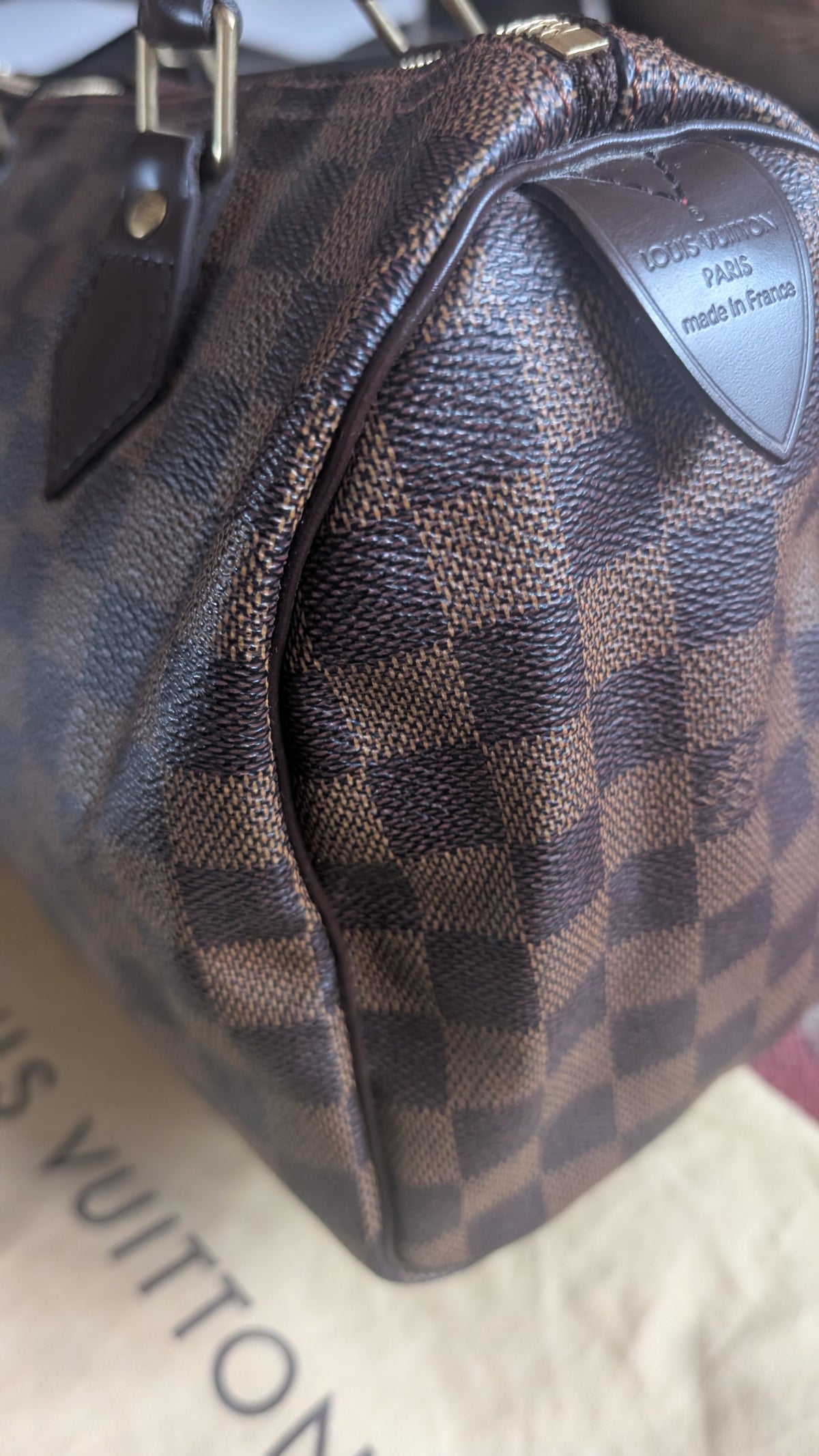 Close-up of a Louis Vuitton handbag in the Damier Ebene canvas pattern, showing the brown and beige checkered design and a leather brand tab on a beige dust bag. Pre-Owned Louis Vuitton Speedy 25 in classic Damier Ebene coated canvas