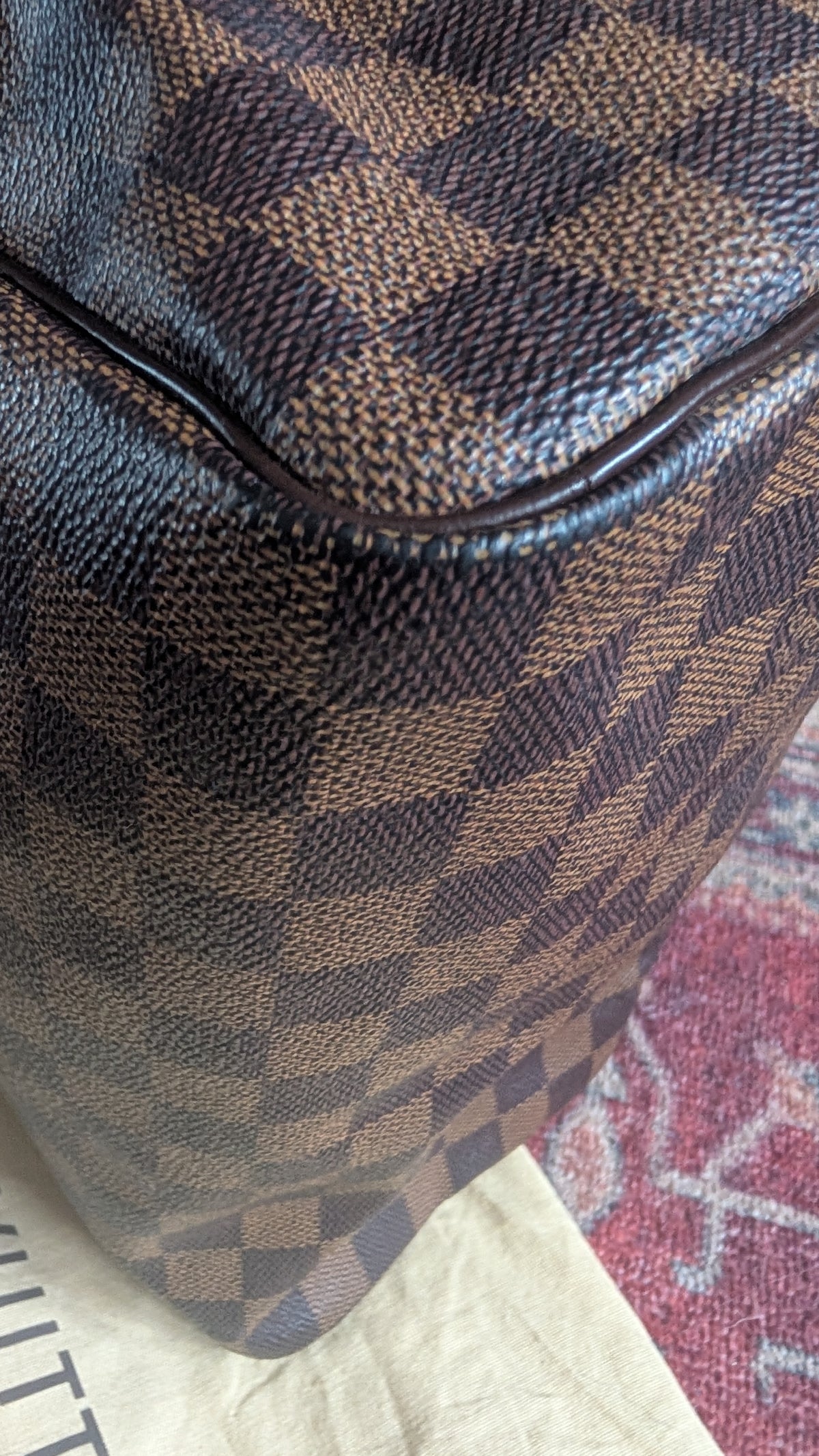 Close-up of a brown and beige checkered patterned bag corner with dark trim, resting on a light surface over a red patterned rug. Pre-Owned Louis Vuitton Speedy 25 in classic Damier Ebene coated canvas