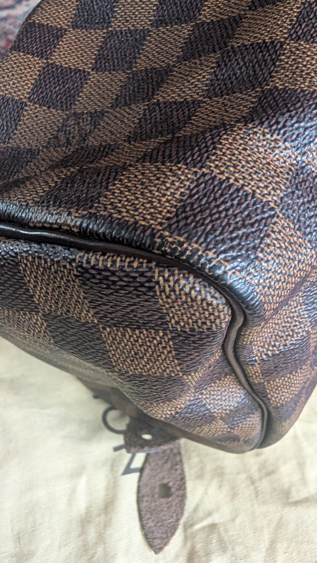 Close-up of a brown and beige checkered canvas bag corner with dark brown leather trim and a small hanging tag, photographed on a light fabric surface. Pre-Owned Louis Vuitton Speedy 25 in classic Damier Ebene coated canvas
