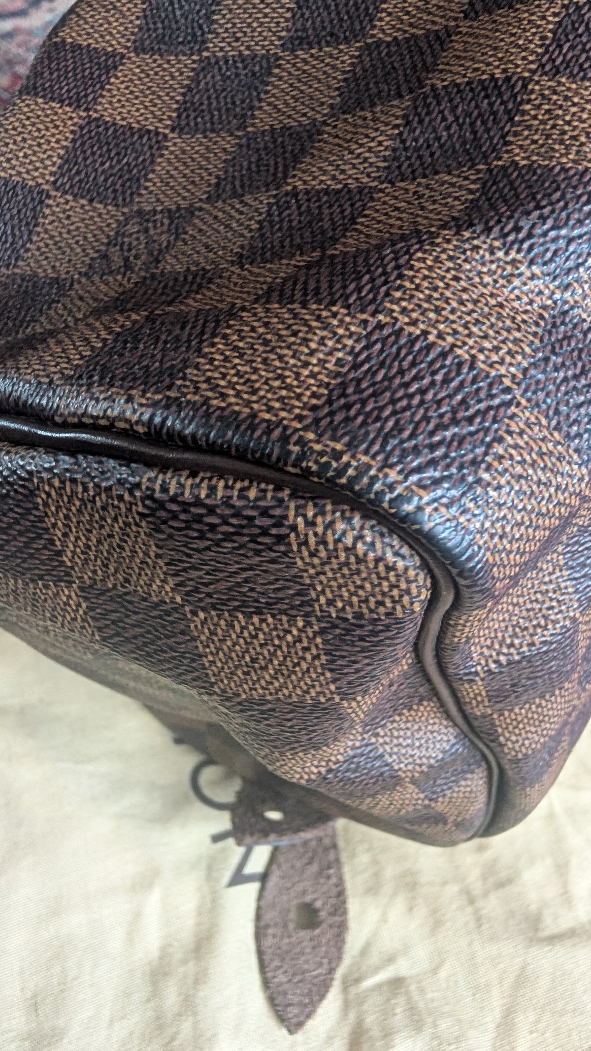 Close-up of a brown and beige checkered canvas bag corner with dark brown leather trim and a small hanging tag, photographed on a light fabric surface. Pre-Owned Louis Vuitton Speedy 25 in classic Damier Ebene coated canvas