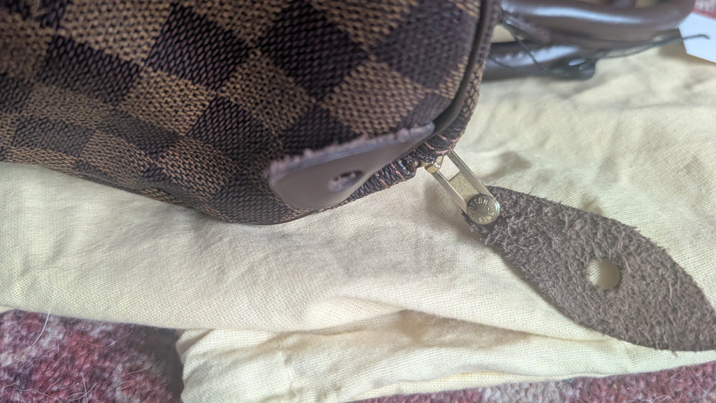 A close-up of a brown and black checkered canvas bag with a brown leather zipper pull, resting on a cream-colored fabric surface. Pre-Owned Louis Vuitton Speedy 25 in classic Damier Ebene coated canvas