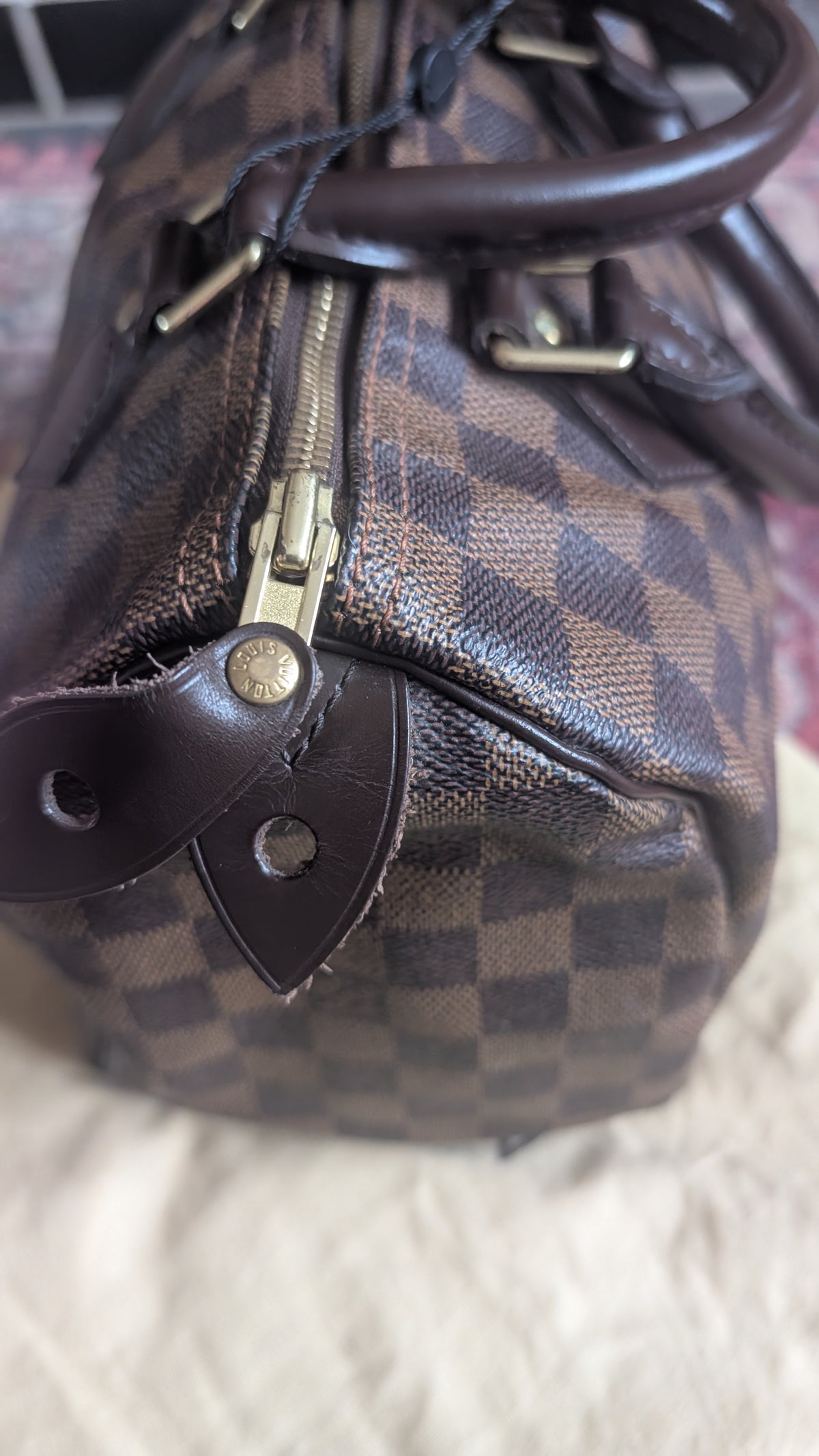 A close-up of a brown checkered canvas handbag with dark brown leather trim, a gold-tone zipper, and metal hardware, resting on a light textured surface. Pre-Owned Louis Vuitton Speedy 25 in classic Damier Ebene coated canvas