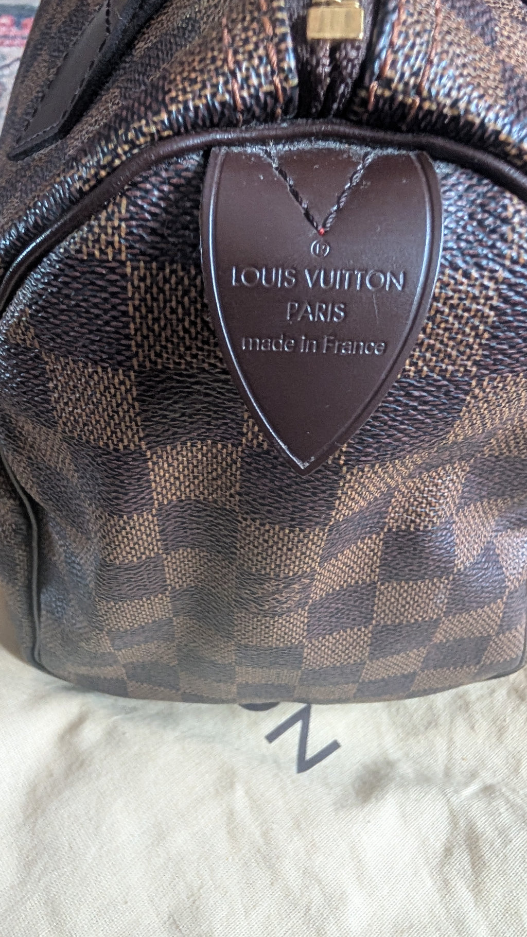 Close-up of a Louis Vuitton handbag in the Damier Ebene check pattern, showing the dark brown leather logo plaque on the front. Pre-Owned Louis Vuitton Speedy 25 in classic Damier Ebene coated canvas