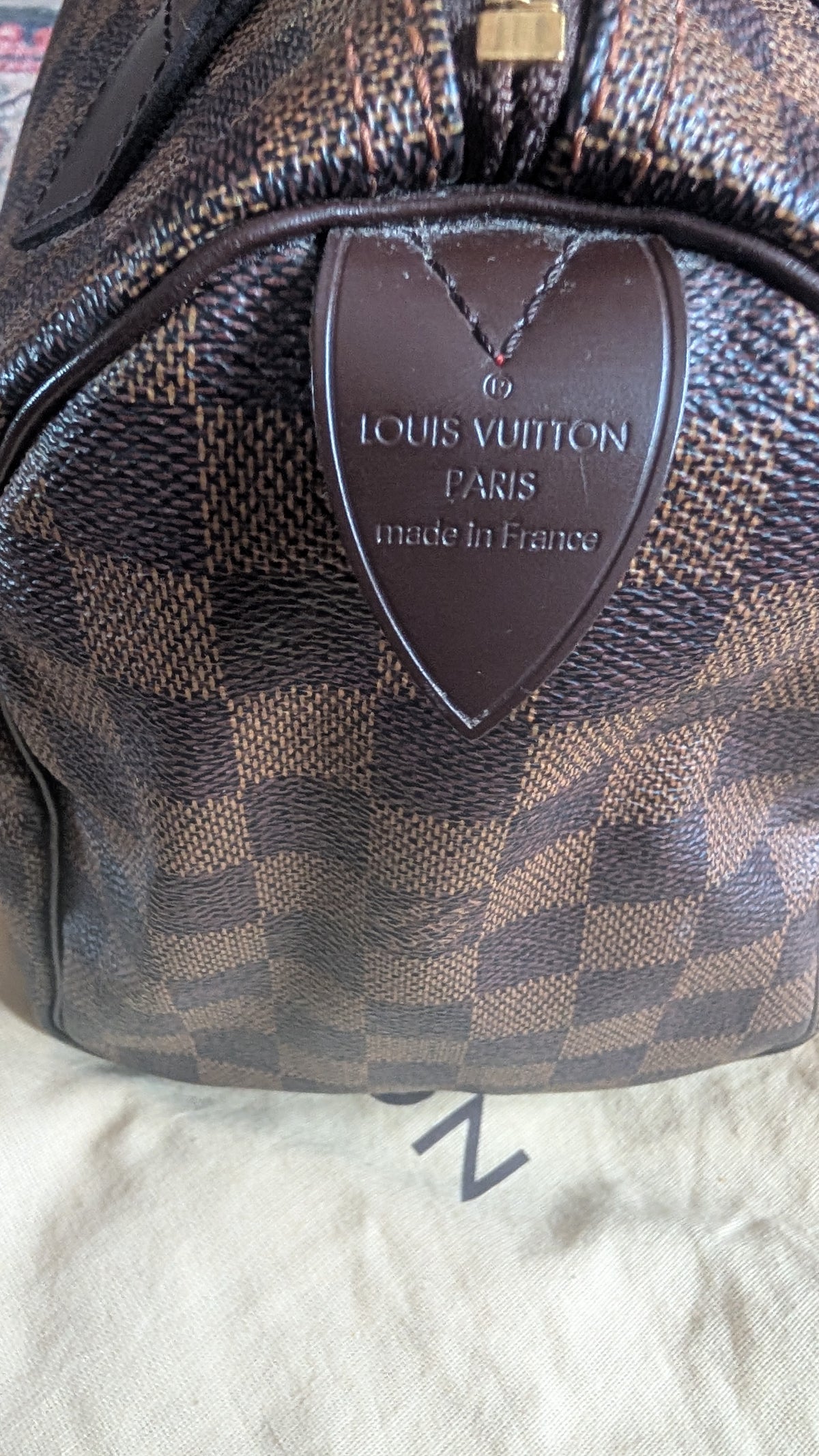 Close-up of a Louis Vuitton handbag in the Damier Ebene check pattern, showing the dark brown leather logo plaque on the front. Pre-Owned Louis Vuitton Speedy 25 in classic Damier Ebene coated canvas