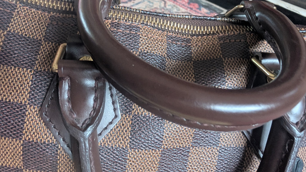 Close-up of a brown leather handbag handle over a brown and beige checkered canvas body with gold-tone hardware. Pre-Owned Louis Vuitton Speedy 25 in classic Damier Ebene coated canvas