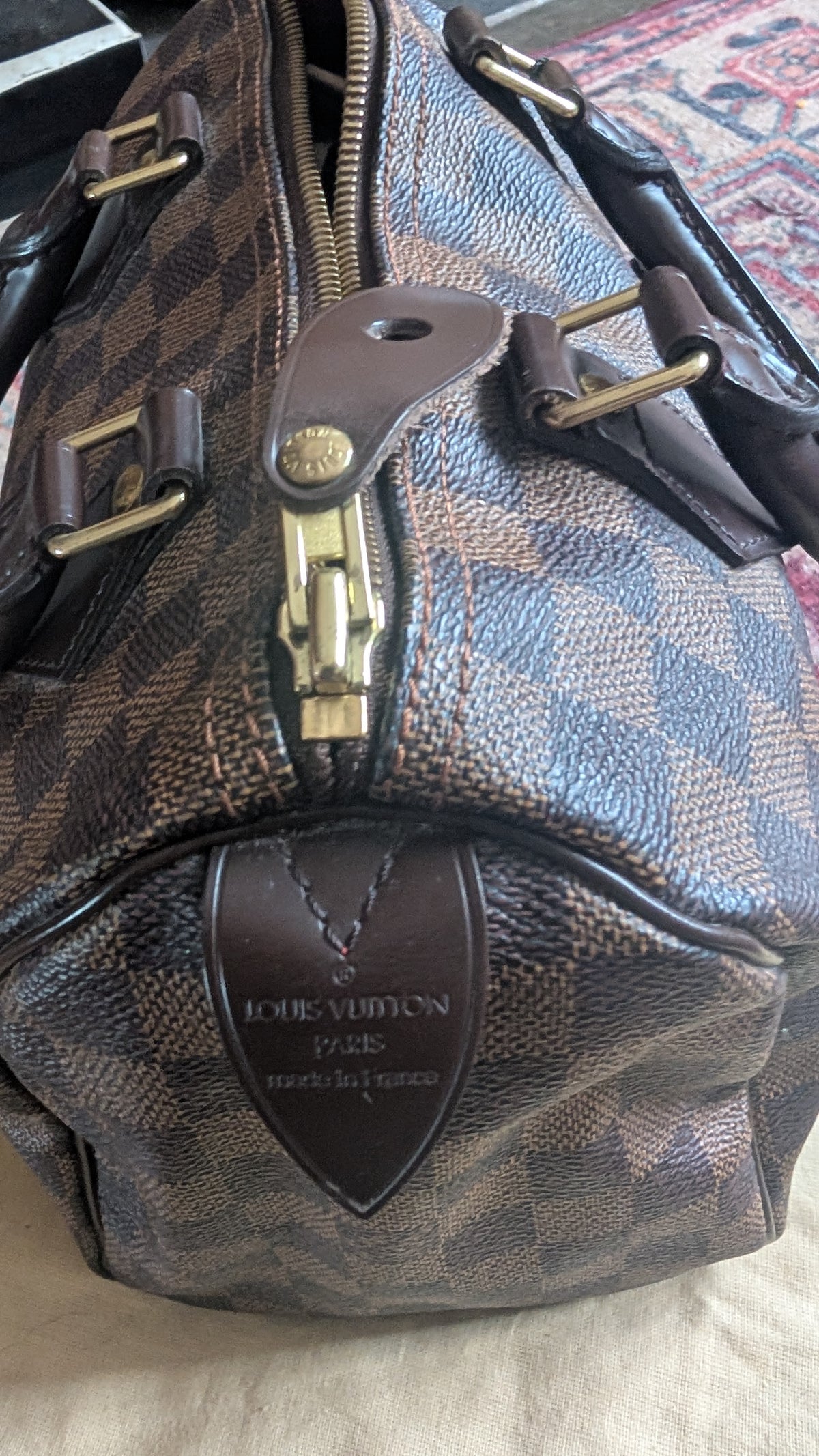 Louis Vuitton Damier canvas handbag with brown leather trim and gold-tone hardware, photographed close up on a light fabric surface. Pre-Owned Louis Vuitton Speedy 25 in classic Damier Ebene coated canvas