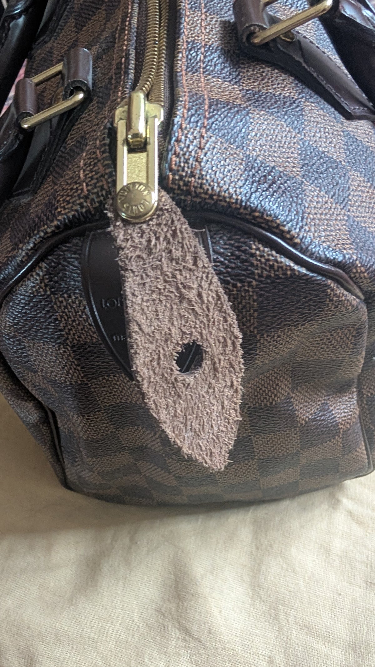 A close-up of a brown and black checkered leather handbag with gold-tone hardware and a fuzzy brown tag, resting on a light beige fabric surface. Pre-Owned Louis Vuitton Speedy 25 in classic Damier Ebene coated canvas