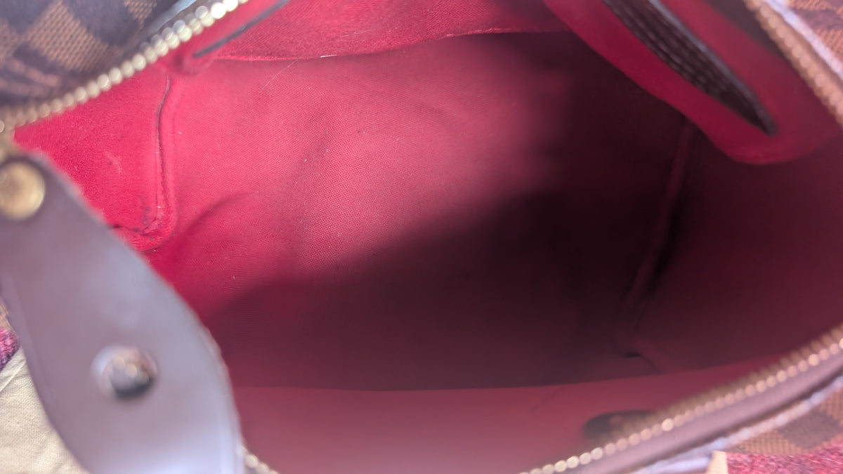 Close-up view of an open handbag showing a dark red or burgundy interior lining, with part of a brown leather strap and a patterned exterior visible at the edges. Pre-Owned Louis Vuitton Speedy 25 in classic Damier Ebene coated canvas
