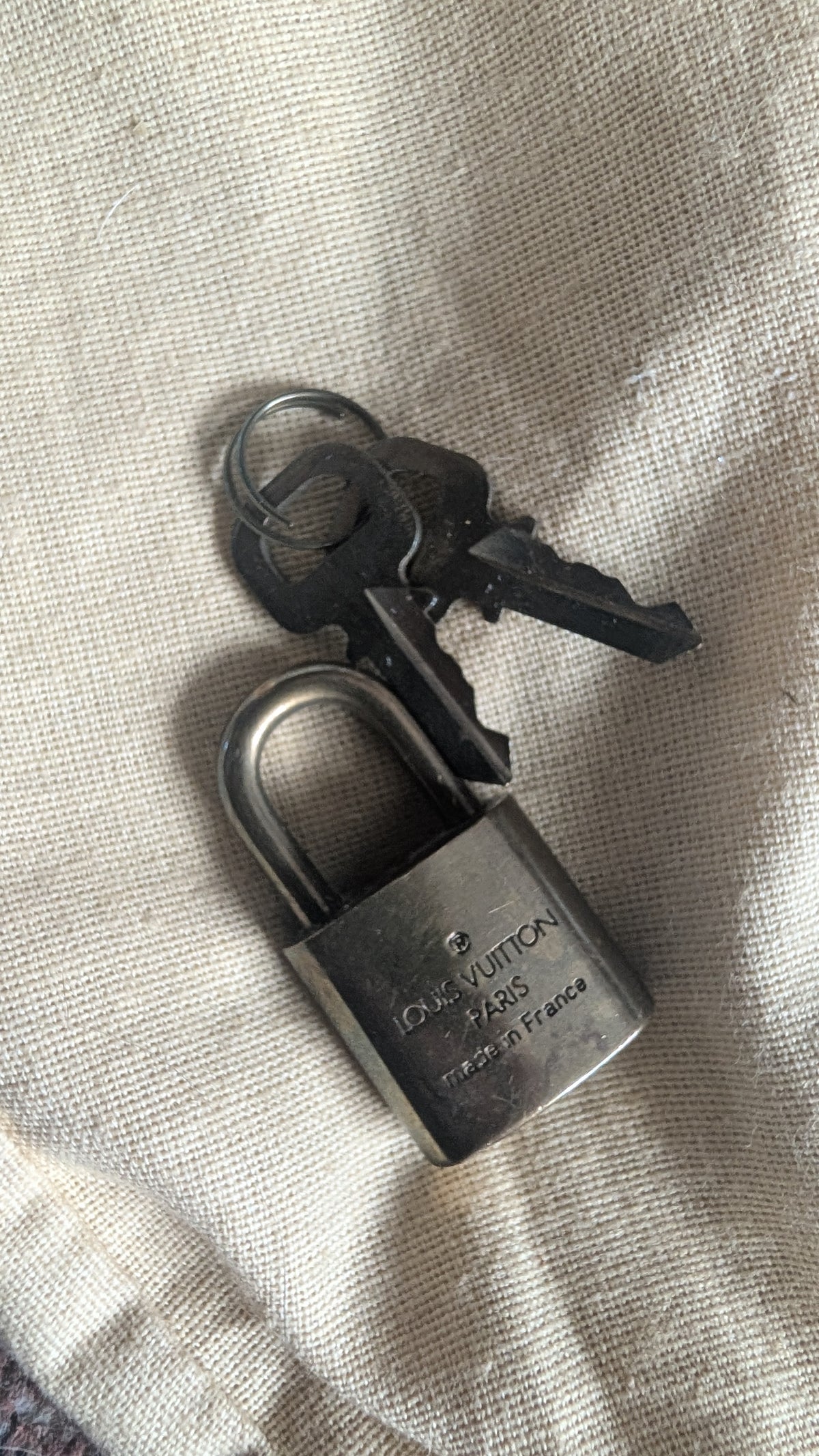A Louis Vuitton padlock with two keys, resting on a textured beige fabric background. The padlock is engraved with 'LOUIS VUITTON PARIS FRANCE'. Pre-Owned Louis Vuitton Speedy 25 in classic Damier Ebene coated canvas
