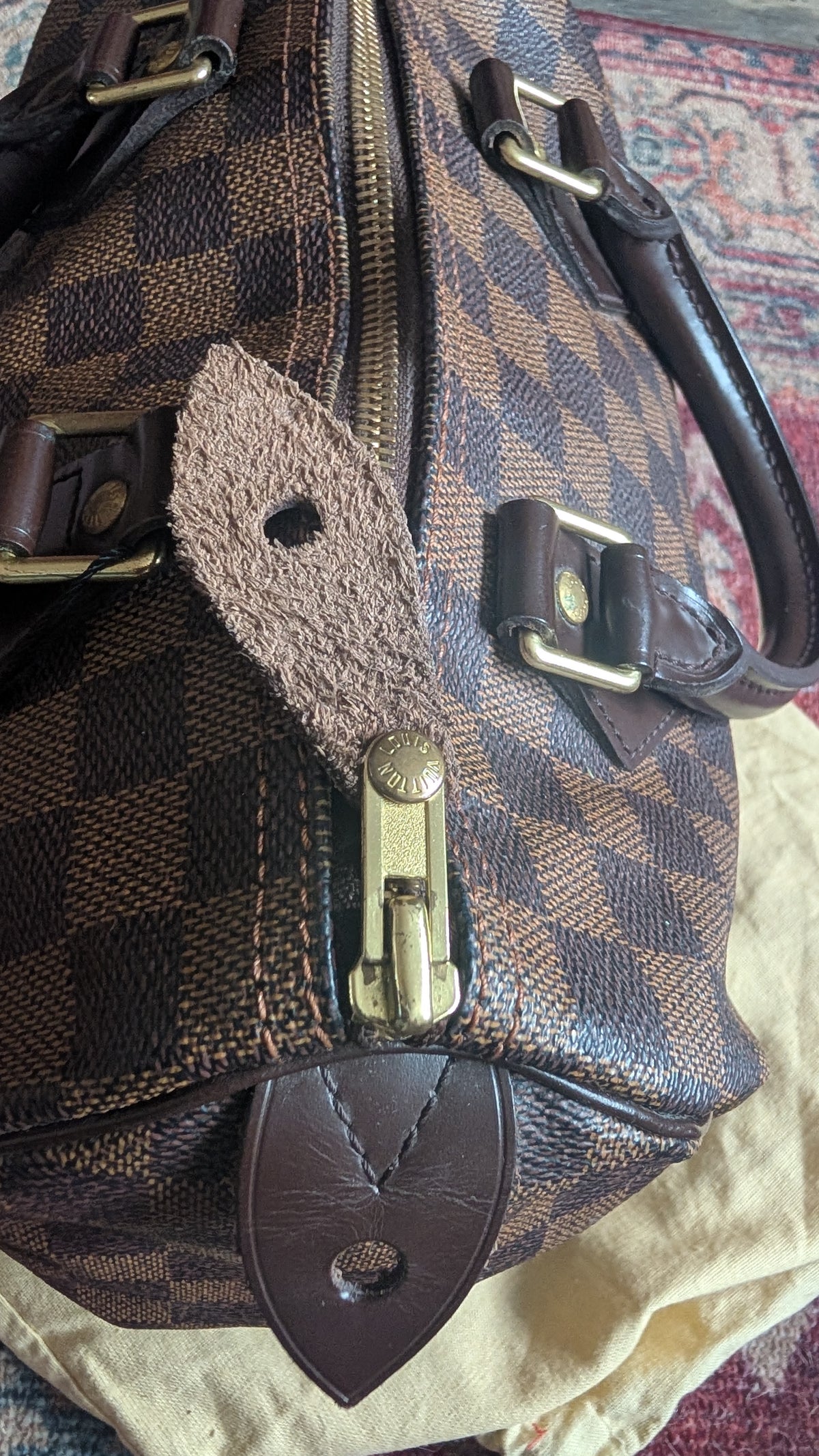 A close-up of a brown and beige checkered leather handbag with gold-tone hardware, a leather strap tab, and a dark brown leather shield-shaped accent, resting on a light beige cloth over a patterned rug. Pre-Owned Louis Vuitton Speedy 25 in classic Damier Ebene coated canvas