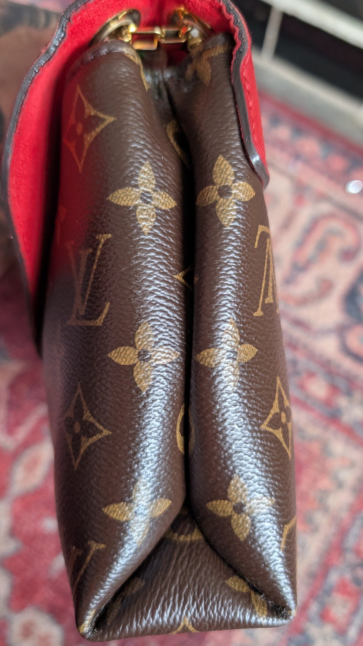 A close-up of a brown Louis Vuitton monogram canvas bag with a red interior lining and gold-tone hardware, resting on a patterned red rug. Pre-Owned Louis Vuitton Pallas Chain Monogram Canvas Shoulder Bag – Cerise Red – M41201
