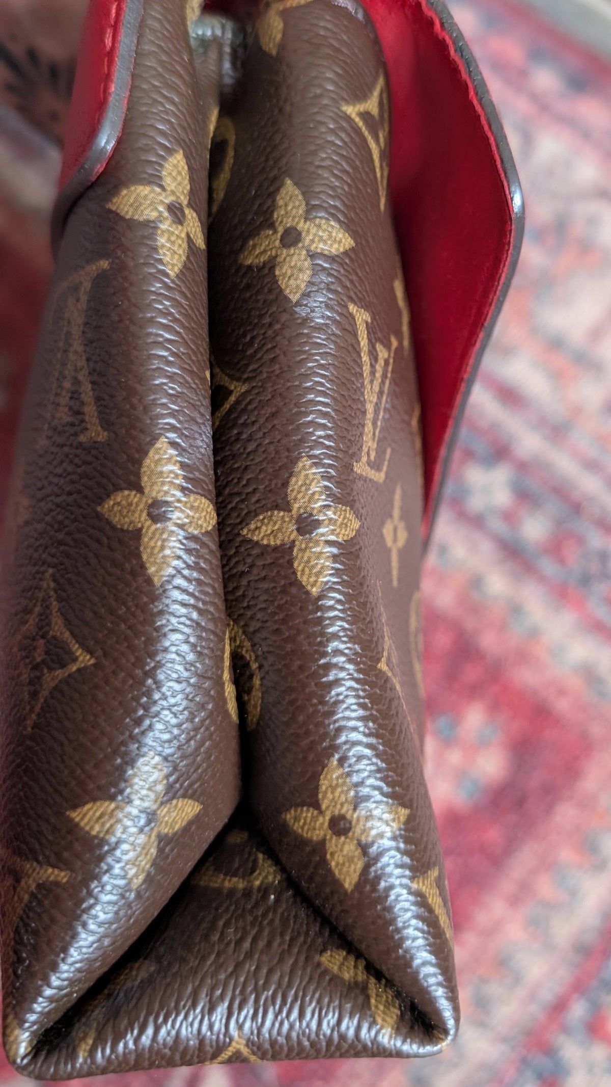 A close-up of a Louis Vuitton monogram handbag corner showing the brown coated canvas with gold-tone LV monogram print and a red interior lining, resting on a patterned red rug. Pre-Owned Louis Vuitton Pallas Chain Monogram Canvas Shoulder Bag – Cerise Red – M41201