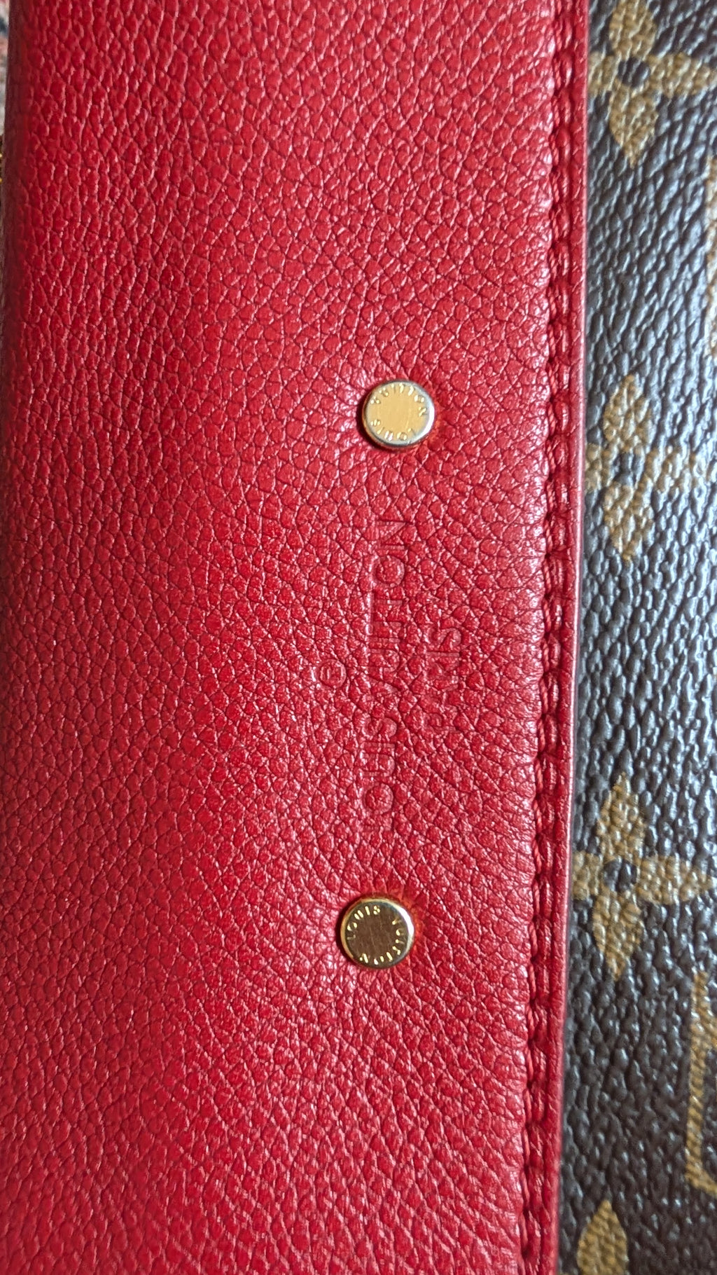 Close-up of a red textured leather wallet or clutch showing two gold-tone metal rivets, with a portion of a brown monogram patterned material visible on the right edge. Pre-Owned Louis Vuitton Pallas Chain Monogram Canvas Shoulder Bag – Cerise Red – M41201
