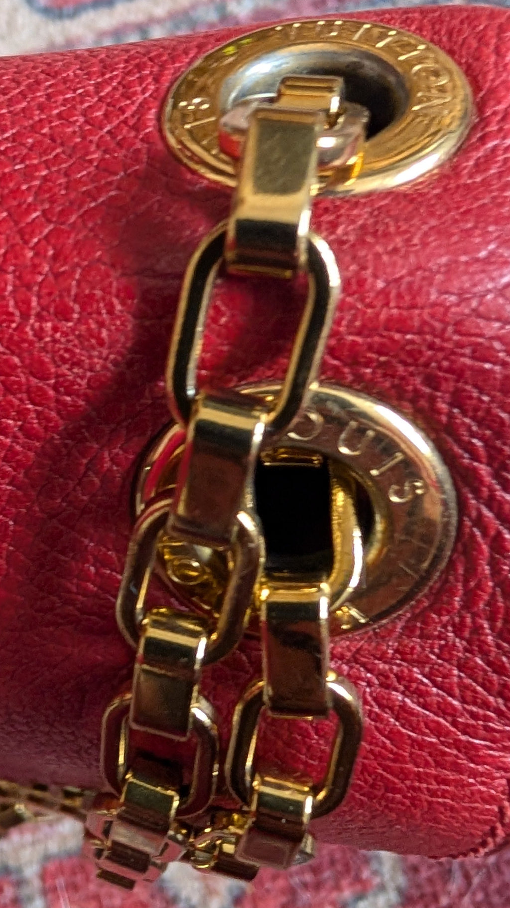 A close-up of a red textured leather handbag with a gold-tone chain strap and circular gold hardware, resting on a patterned rug. Pre-Owned Louis Vuitton Pallas Chain Monogram Canvas Shoulder Bag – Cerise Red – M41201