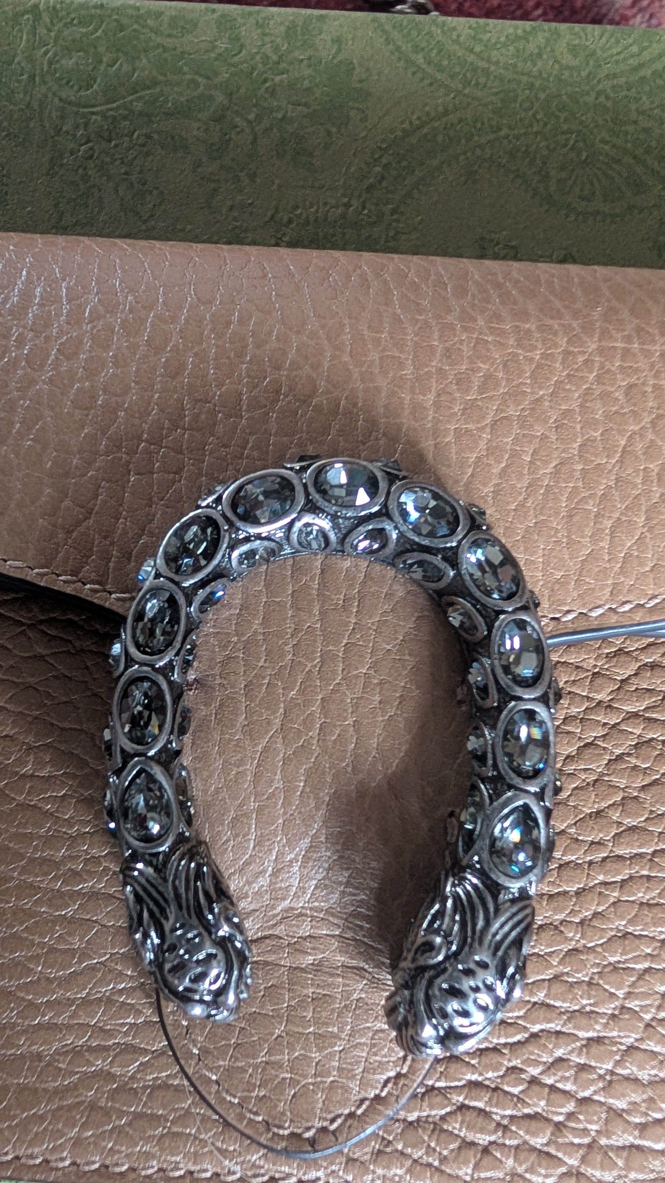 A silver-toned U-shaped metal accessory with ornate scrollwork and circular inset details, displayed on a brown textured leather surface. Pre-Owned Gucci Dionysus Super Mini Crystal Tiger Head Chain Bag – Beige Leather