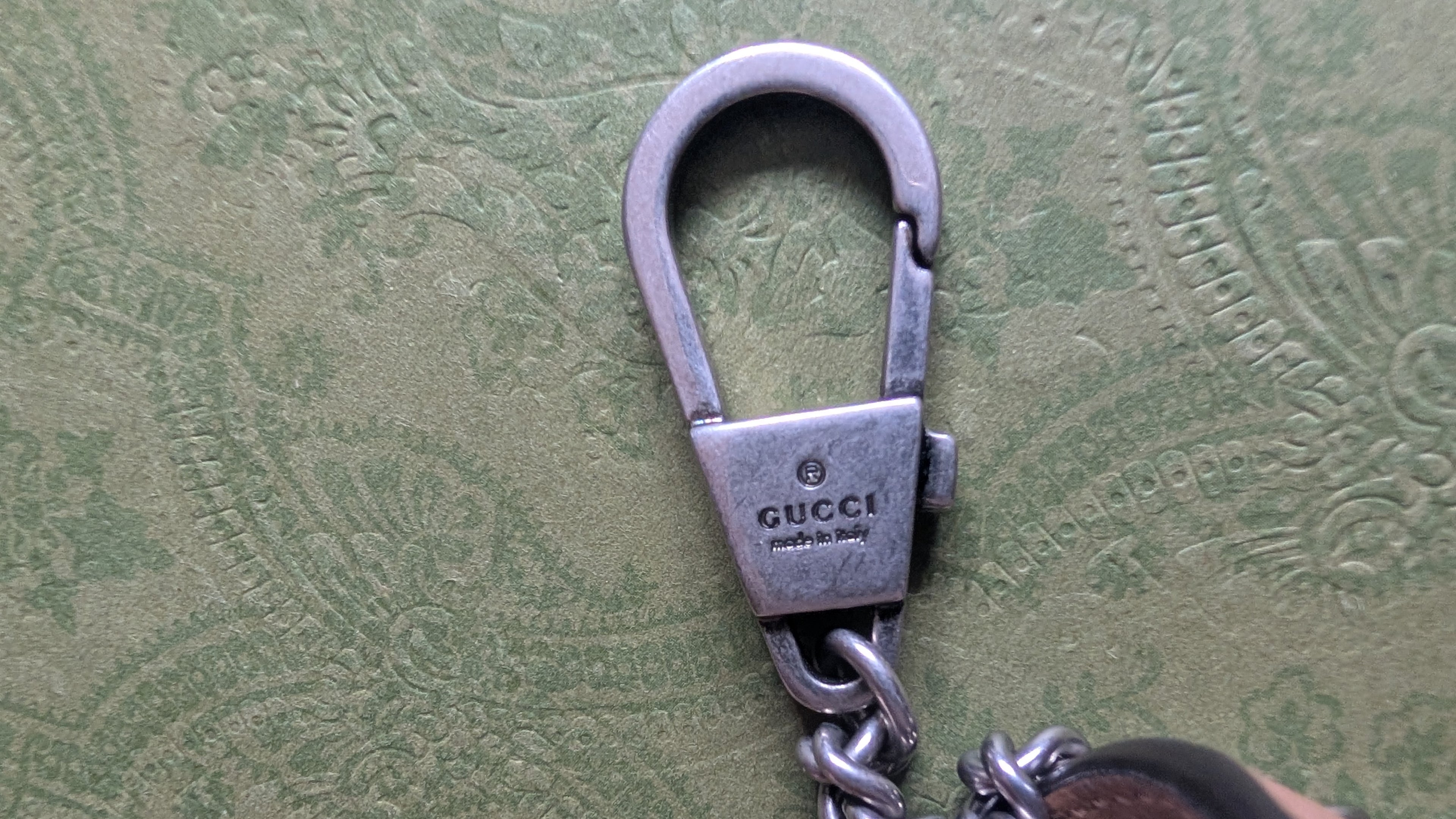 A close-up of a silver-toned metal Gucci keychain clasp with a chain, resting on a textured olive green leather surface. The Gucci brand name is clearly engraved on the clasp. Pre-Owned Gucci Dionysus Super Mini Crystal Tiger Head Chain Bag – Beige Leather