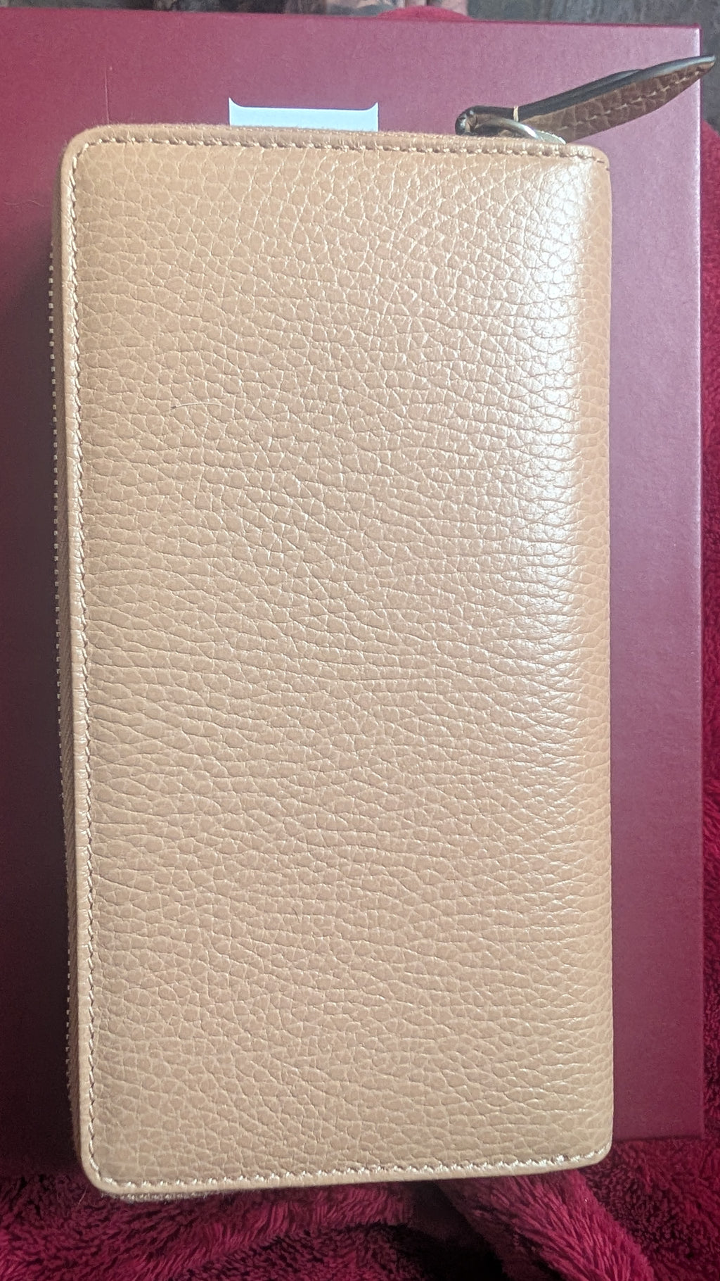 A beige pebbled leather zip-around wallet or card holder, shown closed with a metal zipper pull visible at the top, resting against a dark red background. Pre-Owned Gucci Interlocking G Zip-Around Continental Purse in Nude/Taupe Leather