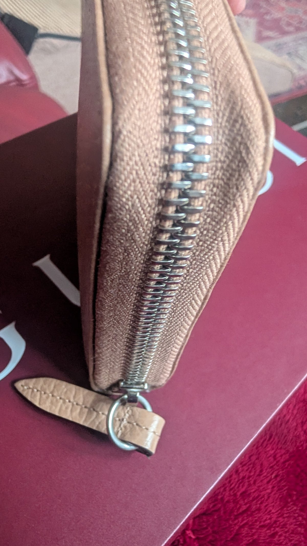A close-up of a textured beige leather zip-around pouch with a silver metal zipper and a matching leather pull tab, resting on a dark red surface. Pre-Owned Gucci Interlocking G Zip-Around Continental Purse in Nude/Taupe Leather