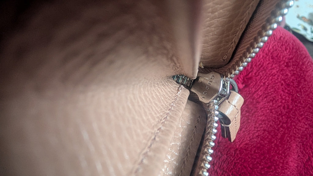 Close-up of a beige pebbled leather zipper pull with a silver metal zipper and a small leather tag, next to a bright pink textured fabric. Pre-Owned Gucci Interlocking G Zip-Around Continental Purse in Nude/Taupe Leather
