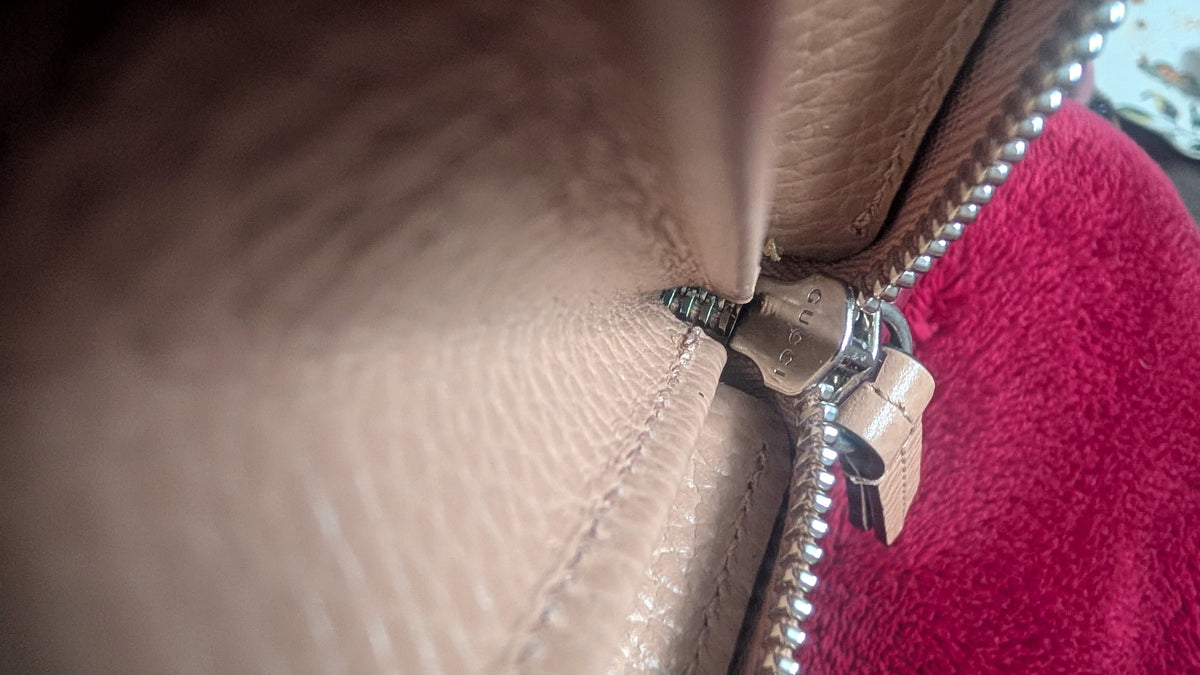 Close-up of a beige pebbled leather zipper pull with a silver metal zipper and a small leather tag, next to a bright pink textured fabric. Pre-Owned Gucci Interlocking G Zip-Around Continental Purse in Nude/Taupe Leather