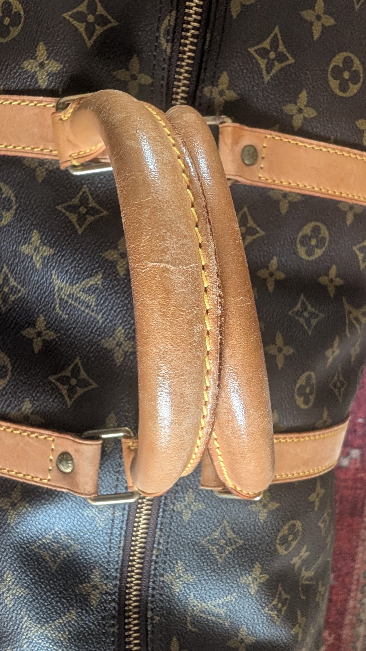 Close-up of a Louis Vuitton monogram duffel bag with a tan leather handle and matching leather trim on a dark brown background. Pre-Owned Louis Vuitton Keepall Bandoulière 60 Monogram Travel Bag